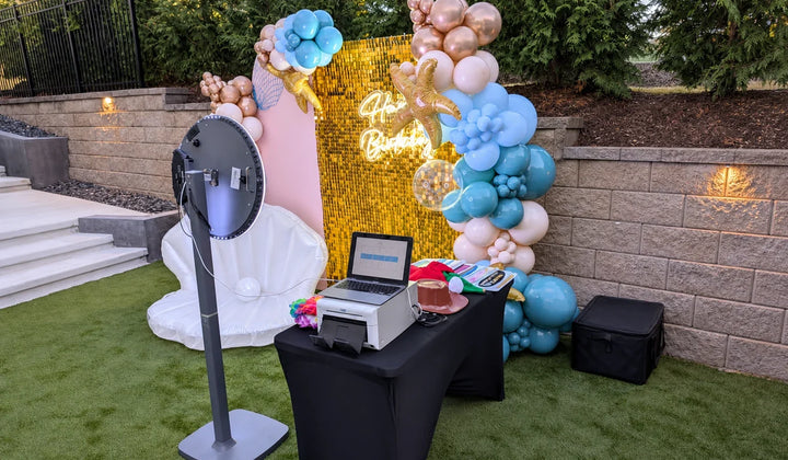 Photo booth setup with printer, laptop, gold backdrop, and balloon decor at an outdoor event