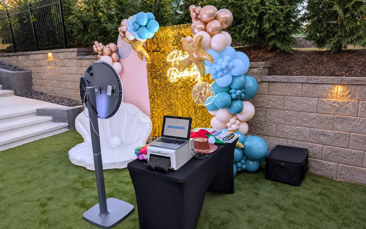 Photo booth setup with printer, laptop, gold backdrop, and balloon decor at an outdoor event