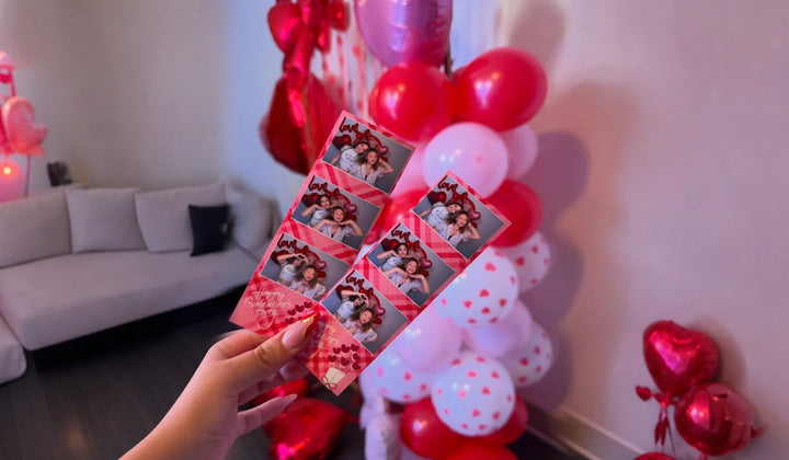 Hand holding two printed photo booth strips in front of Valentine event balloons