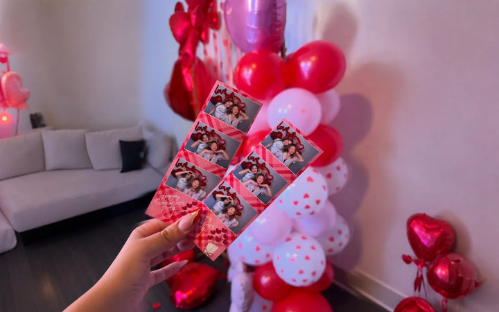 Hand holding two printed photo booth strips in front of Valentine event balloons